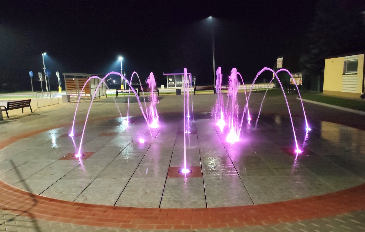 Z fontanny tryskają strumienie wody podświetlone na kolor fioletowy, wieczorne niebo w tle.